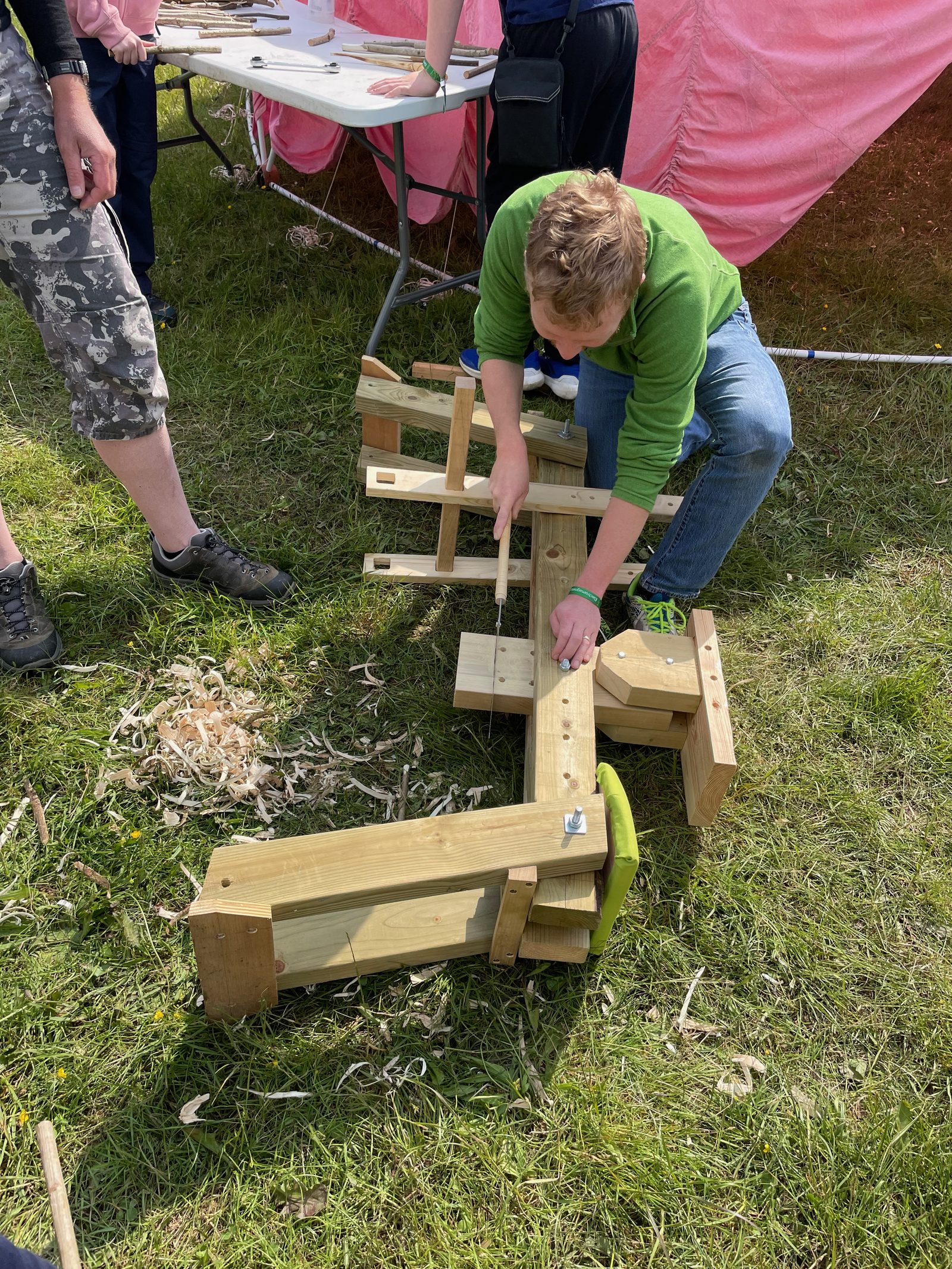 A young person working at a shave horse in front of a pink geodesic dome at EMF 2024, wood shavings scattered on the grass.